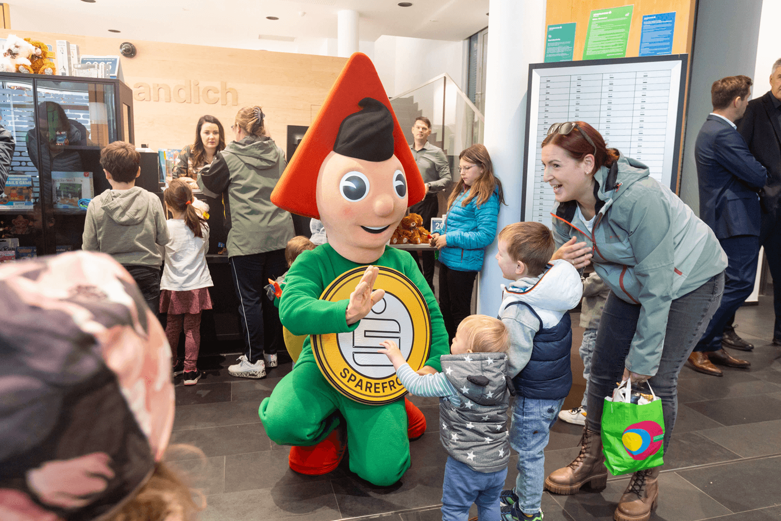 Sparkassen-Maskottchen Sparefroh in einer Filiale mit zwei Kindern und ihrer Mutter beim Abklatschen.