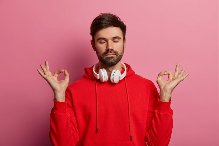 Studio shot of male youngster makes mudra zen gesture, keeps eyes shut, wears headphones around neck, meditates and breathes deeply, listens relaxing music, has red sweatshirt, poses over rosy wall