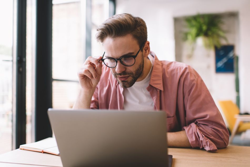 Skilled hipster guy in optical eyewear browsing informative website on digital laptop technology using public internet connection in coworking space,Caucasian male programmer in glasses installing app