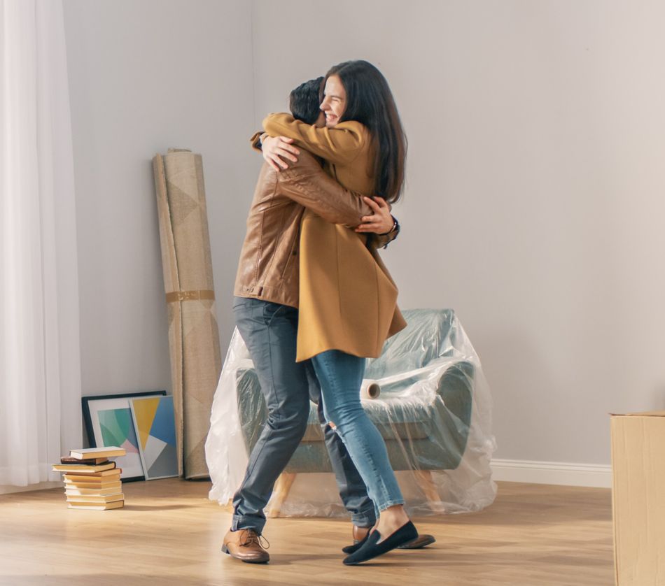 Happy and Excited Young Couple Look Around In Wonder at their Newly Purchased / Rented Apartment. Beautiful People Happily Embracing. Big Bright Modern Home with Cardboard Boxes Ready to Unpack.