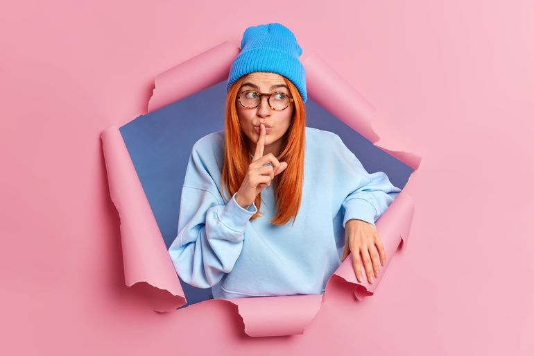 Mysterious redhead woman makes silence gesture asks not to tell her secret wears blue hat and sweate