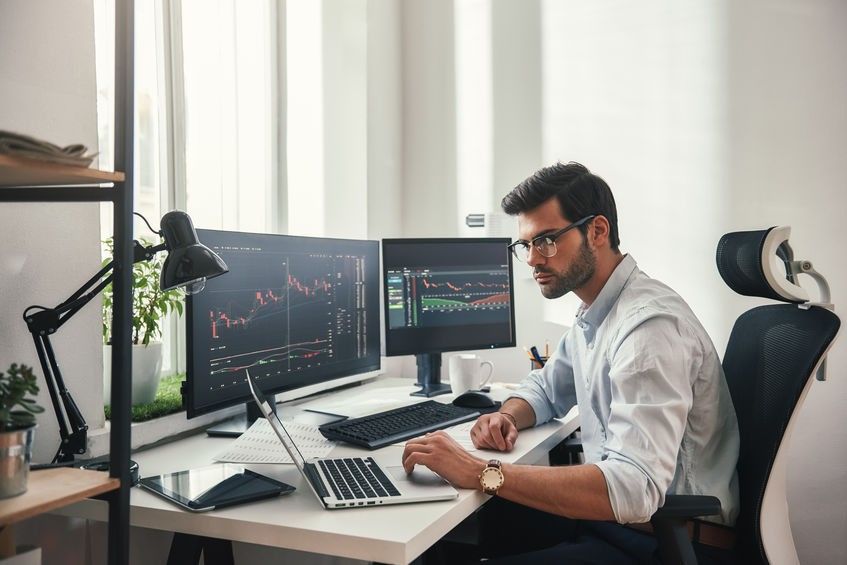 Busy working day. Young bearded trader in eyeglasses working with laptop while sitting in his modern office in front of computer screens with trading charts.