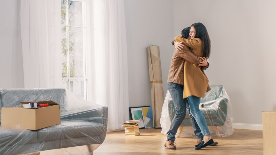 Happy and Excited Young Couple Look Around In Wonder at their Newly Purchased / Rented Apartment. Beautiful People Happily Embracing. Big Bright Modern Home with Cardboard Boxes Ready to Unpack.