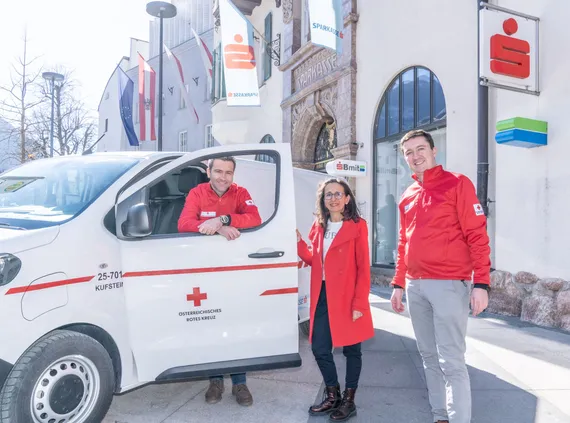 drei Personen posieren vor dem Roten Kreuz Auto