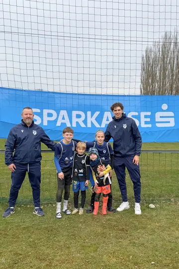 Zwei Männer in Trainingsanzügen und vier Kinder in Fußballbekleidung auf dem Spielfeld