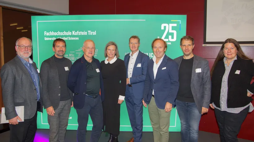 8 Personen stehen vor einem Fachhochschule Kufstein Aufsteller und lächeln in die Kamera