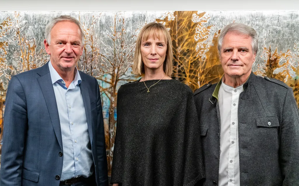zwei Herren, eine Dame blicken entspannt in die Kamera, hinter ihnen ein Gemälde