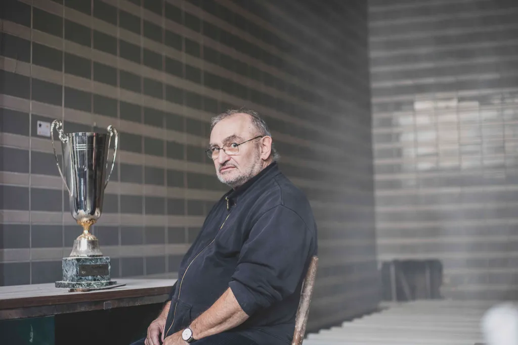 Herr sitzt auf Stuhl, am Tisch neben ihm ein Pokal, er blickt in die Kamera