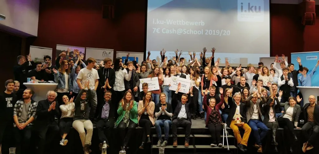Gruppenfoto mit den Teilnehmenden des i.ku-Wettbewerbs auf der Bühne, manche strecken die Hände in die Höhe