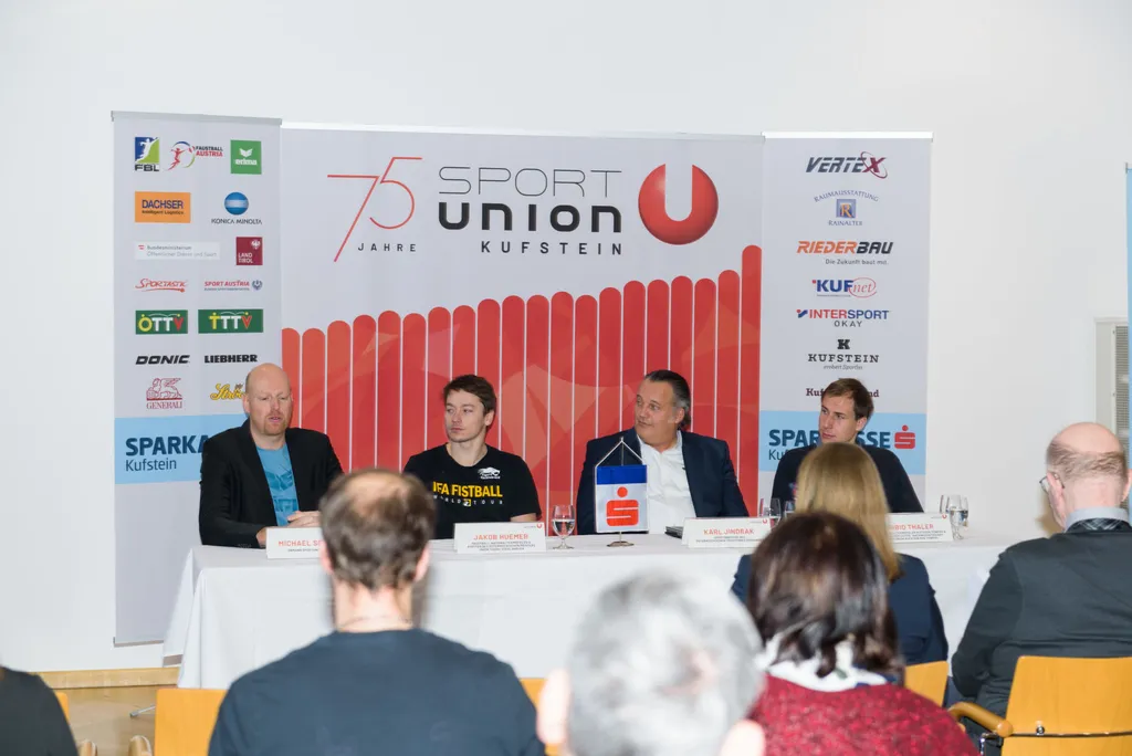 vier Herren sitzen vorne an einem Tisch und reden zu den Zuhörenden, hinter ihnen das Logo der Sport Union Kufstein