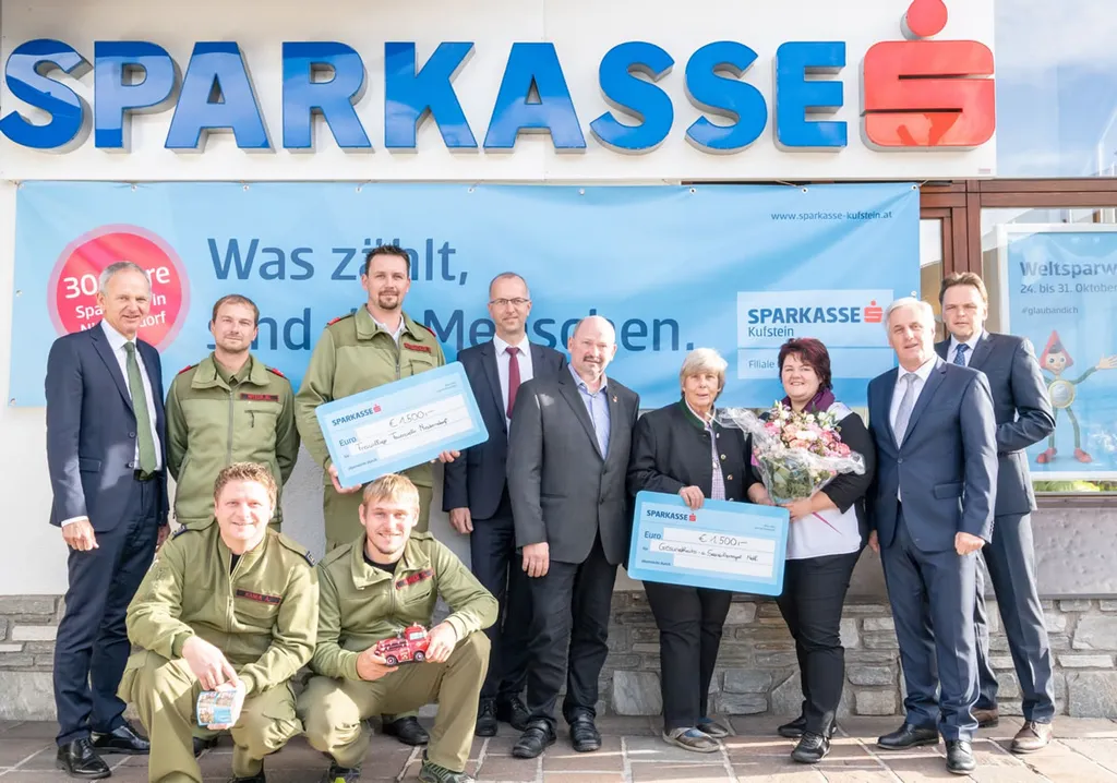 ein Feuerwehrmann und eine Frau halten eine Spendentafel in der Hand- sieben Personen stehen nebenan und zwei Feuerwehrmänner sind hockend im Vordergrund