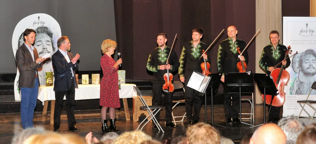 auf der Bühne stehen vier Männer mit Geige in der Hand, eine Frau mit Mikrofon und zwei klatschende Männer