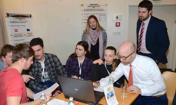sechs Personen sitzen bei Tisch und zwei Personen stehen im Hintergrund 