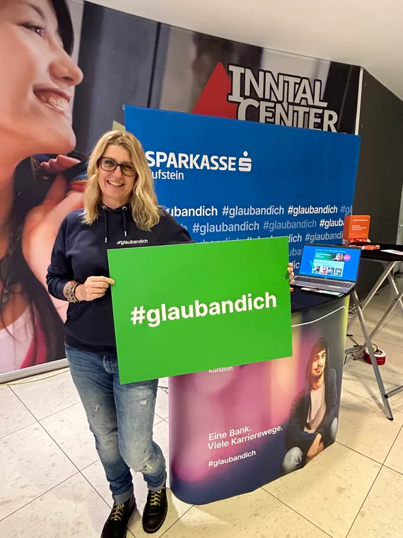 eine Dame steht vor einem Sparkasse Kufstein Messestand und hält ein grünes Schild mit #glaubandich in den Händen