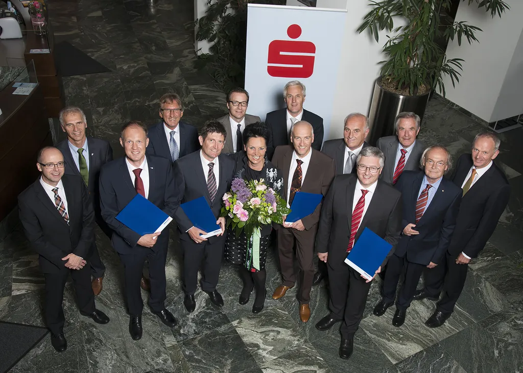 Männer mit Auszeichnung in der Hand und eine Frau mit Blumen in der Hand schauen lachend hinauf zu in die Kamera