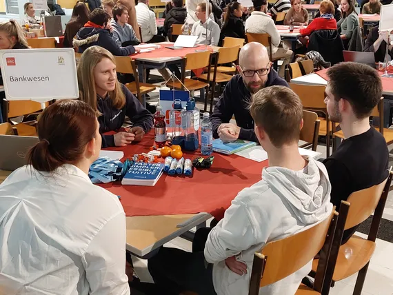 5 Personen sitzen um einen Tisch, die Erwachsenen erklären den Jugendlichen das Thema Bankwesen