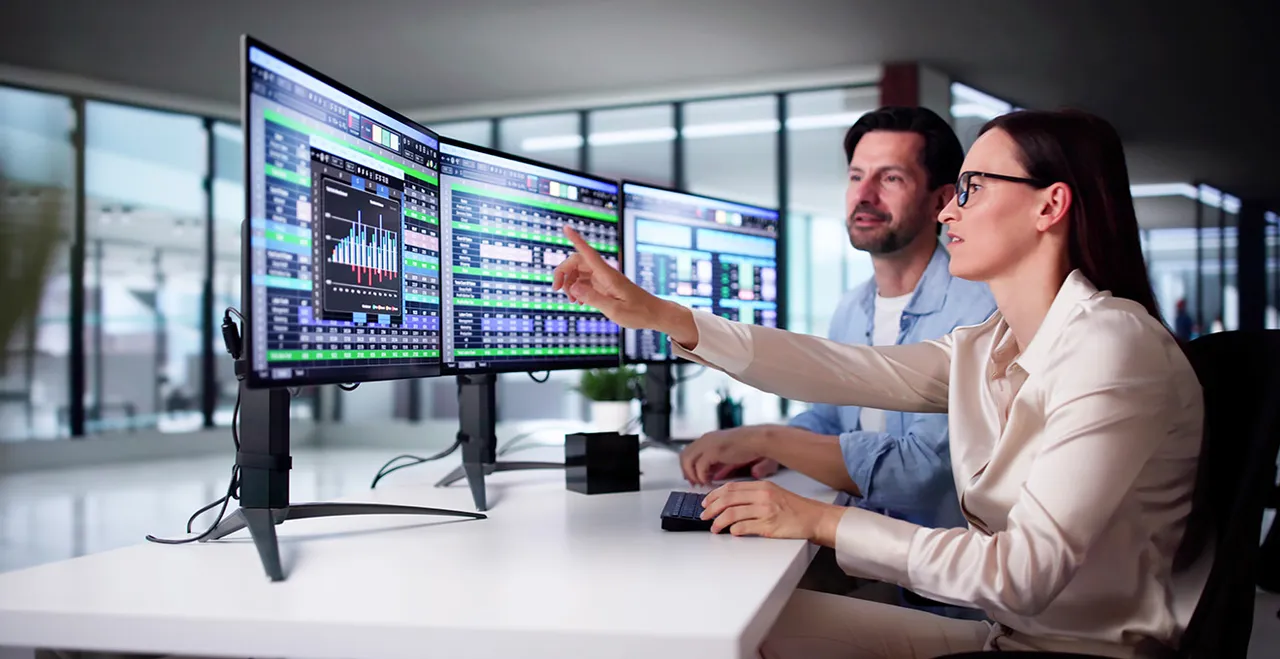 Ein Mann und eine Frau vor drei Monitoren mit Charts