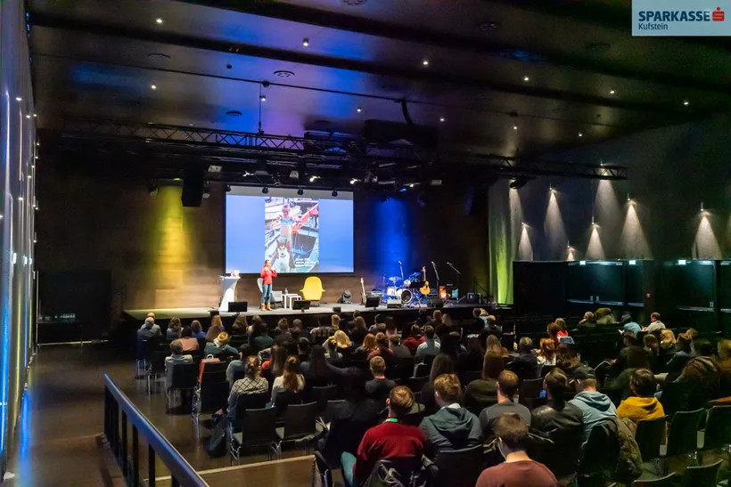 Saal mit zuhörenden Person und einer Person auf der Bühne