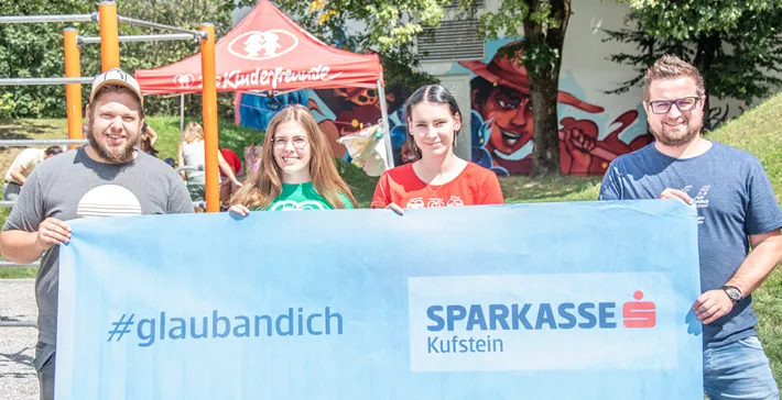 vier Personen halten im Freien ein #glaubandich Banner in die Luft und lachen in die Kamera