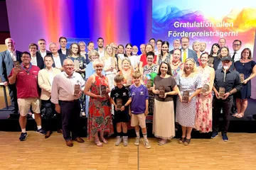 Gruppenfoto von Menschen unterschiedlichen Alters auf einer Bühne vor einer Projektion mit dem Text 'Gratulation allen Förderpreisträgern