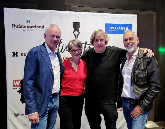 vier Personen posieren Arm in Arm vor einem Aufsteller