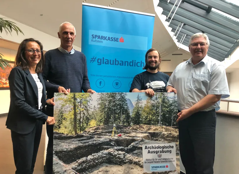 4 Personen stehen vor dem Sparkasse Kufstein Banner und lächeln und halten ein Foto der archäologischen Ausgrabungen in den Händen