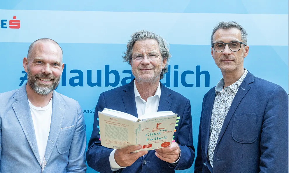drei Herren posieren vor einem #glaubandich Aufsteller, der Herr in der Mitte hält ein Buch