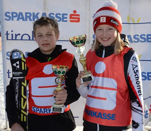 ein Mädchen und ein Junge lachen mit Skianzug und Pokal in der Hand in die Kamera
