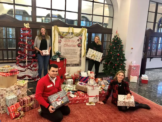 2 Personen stehen, 2 Personen sitzen umgeben von Weihnachtsdekoration und halten Geschenke in den Händen
