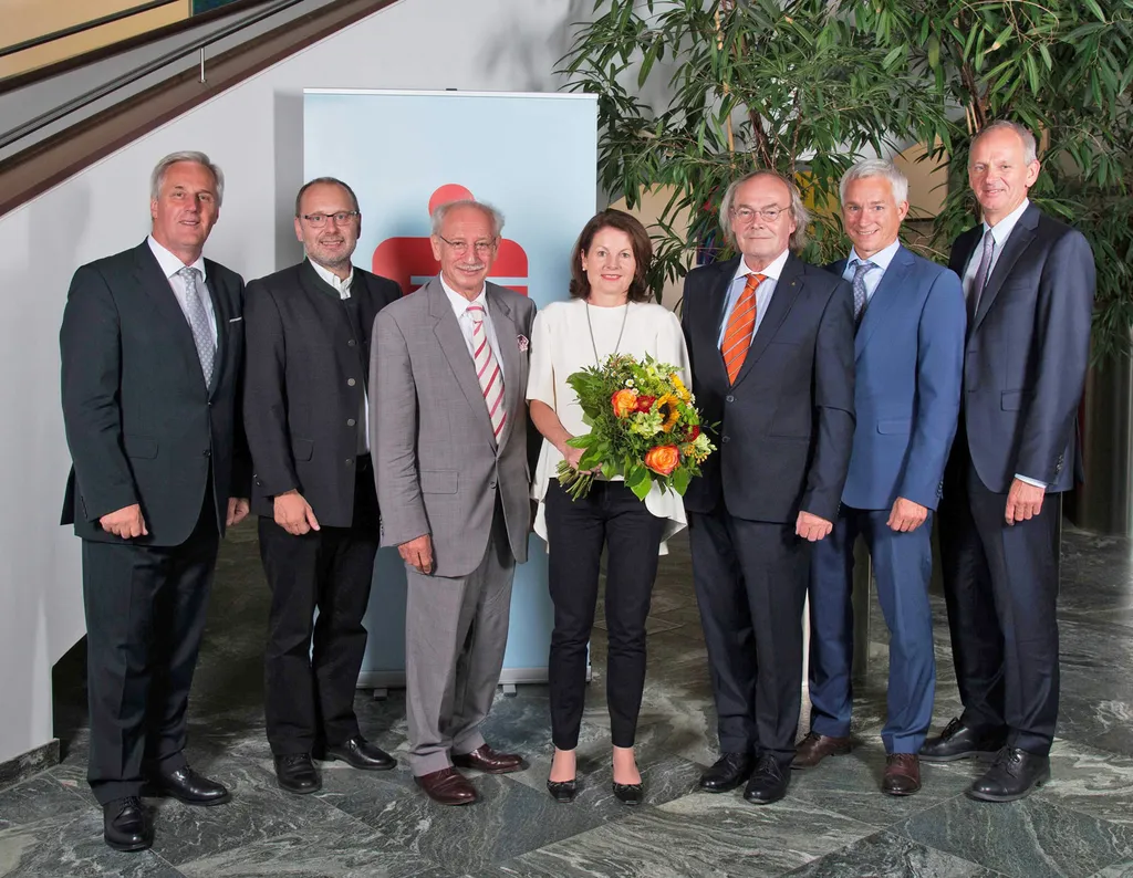 sechs Männer und eine Frau mit Blumen in der Hand lachen stehend in die Kamera