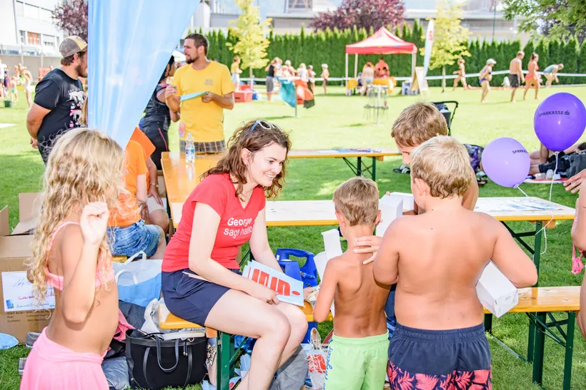 Eine Dame sitzt auf einer Bank umringt von Kindern beim Spielfest im Freien