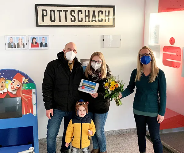 Kundenbetreuerin Elke Hahnekamp (rechts) übergibt den glücklichen Gewinnern Julia und Thomas Ritter ihren Hochzeitsreise-Gutschein in der Sparkassen-Filiale Pottschach. Auch der kleine Lukas freut sich für seine Eltern.