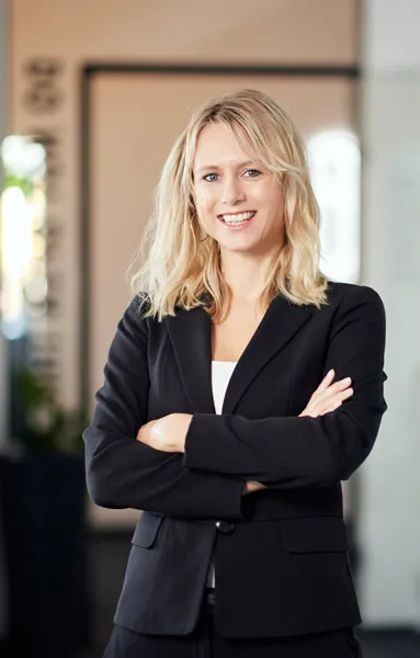 Dame mit blonden Haaren und schwarzem Blazer hat die Arme verschränkt und lächelt in die Kamera