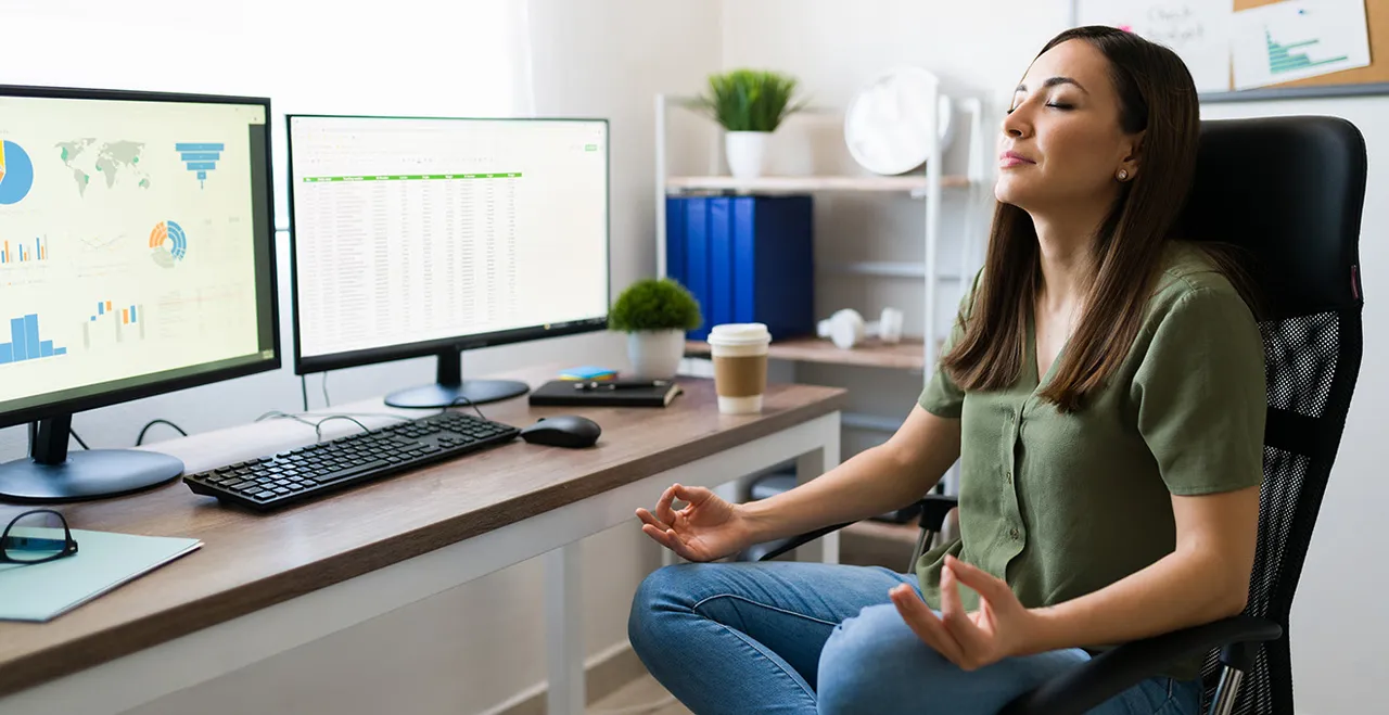 Meditierende Frau auf einem Bürosessel, daneben zwei Monitore mit Charts