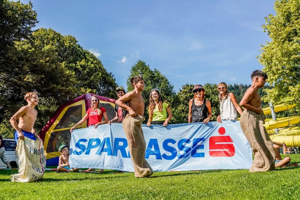 drei Kinder spielen Sackhüpfen und fünf Kinder halten das Sparkasse Transparent im Hintergrund 