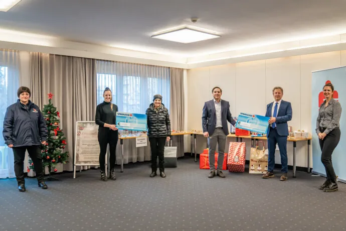 6 Personen stehen im Halbkreis und lächeln in die Kamera, der Raum ist weihnachtlich geschmückt und sie halten zwei Schecks in den Händen