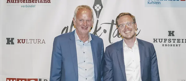 2 Herren in Anzug stehen vor einem Werbebanner und lächeln