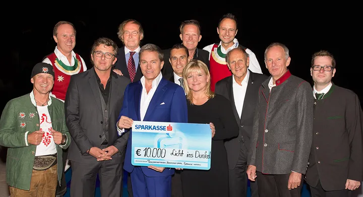 ein Mann und eine Frau halten Spendentafel in der Hand und viele Personen stehen im Hintergrund