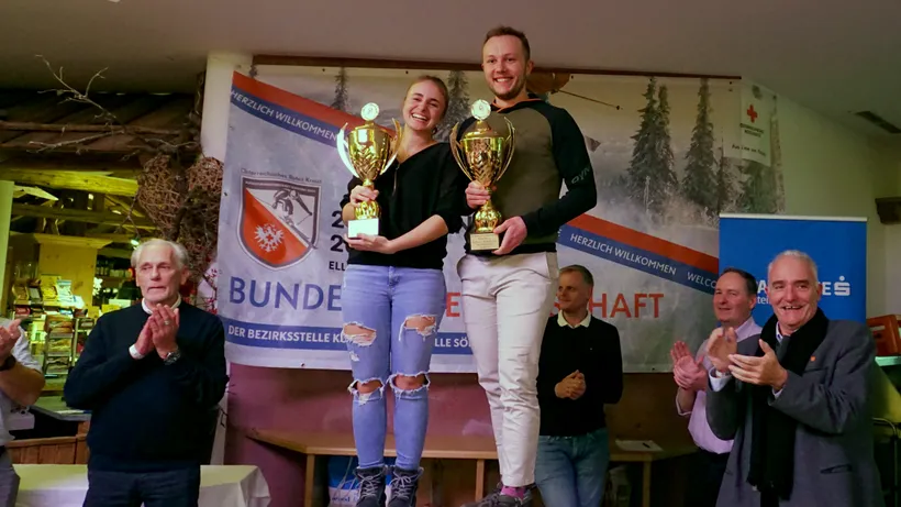 eine junge Frau und ein junger Mann halten jeweils einen Pokal während die anderen Personen applaudieren