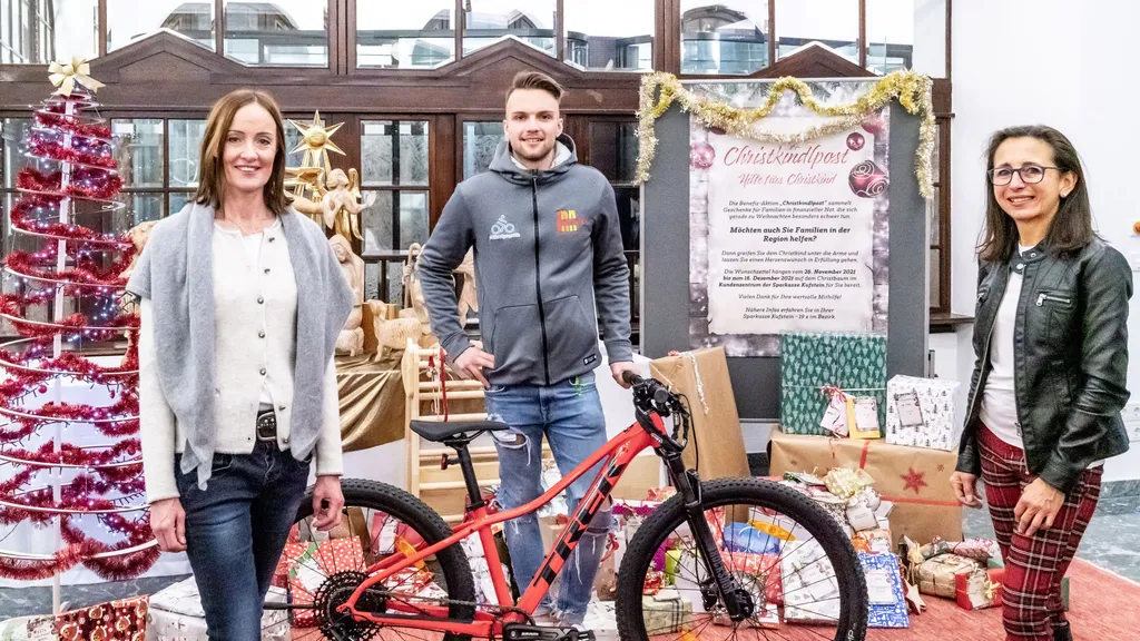 3 Personen lächeln in die Kamera vor weihnachtlicher Dekoration bei Geschenkübergabe