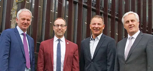 4 Herren in Anzug stehen vor einem Gebäude und lächeln in die Kamera