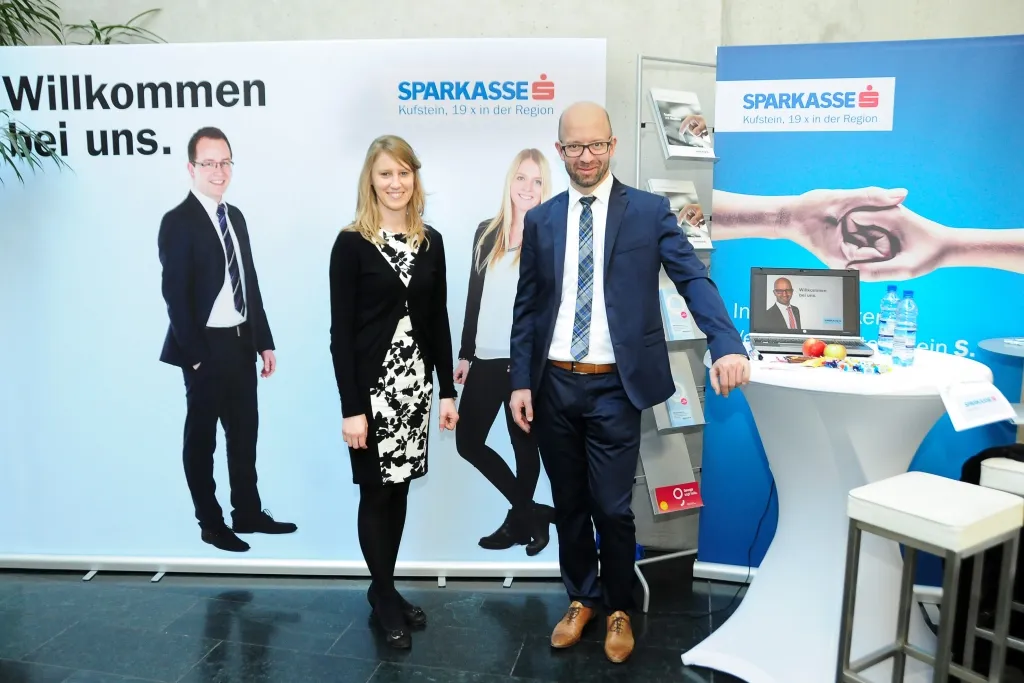 ein Mann und eine Frau stehen vor einem Sparkasse Plakat bei einem Stehtisch