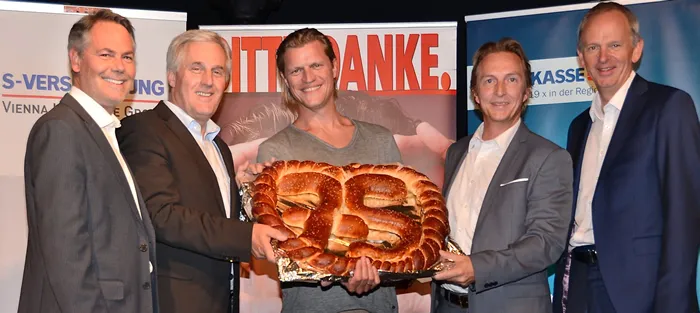 drei Männer halten gemeinsam eine Brezel in der Hand und zwei Männer stehen nebenan