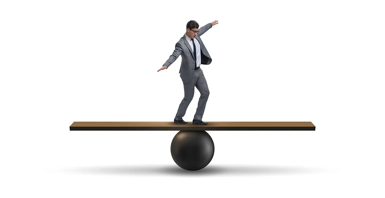 Mann im Anzug balanciert auf Brett, das sich auf einer Kugel befindet