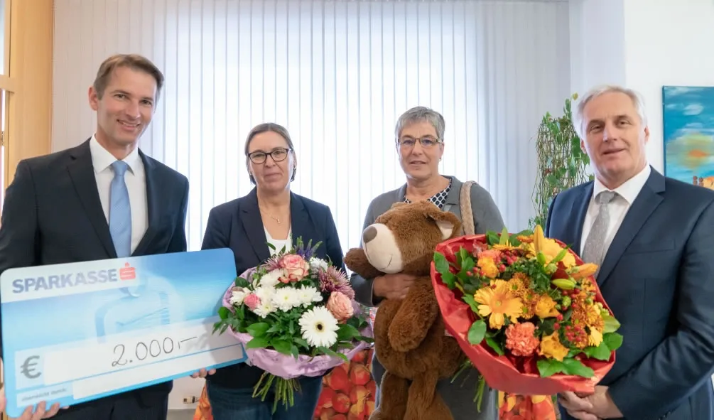ein Mann hält Spendentafel in der Hand, eine Frau hält Blumen in der Hand, nebenan eine Frau mit Teddybär in der Hand und ein Mann