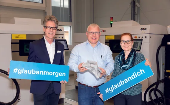 Zwei Männder und eine Frau stehen vor einem Metall-3D-Drucker.