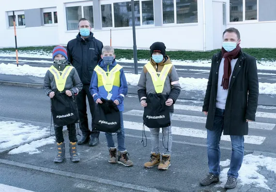 drei Kinder mit Maske am Schulweg halten ein #glaubandich Sackerl in den Händen, daneben zwei Herren ebenfalls mit Marke